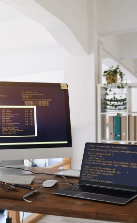 Background image of black and orange programming code on computer screen and laptop in contemporary office interior, copy space